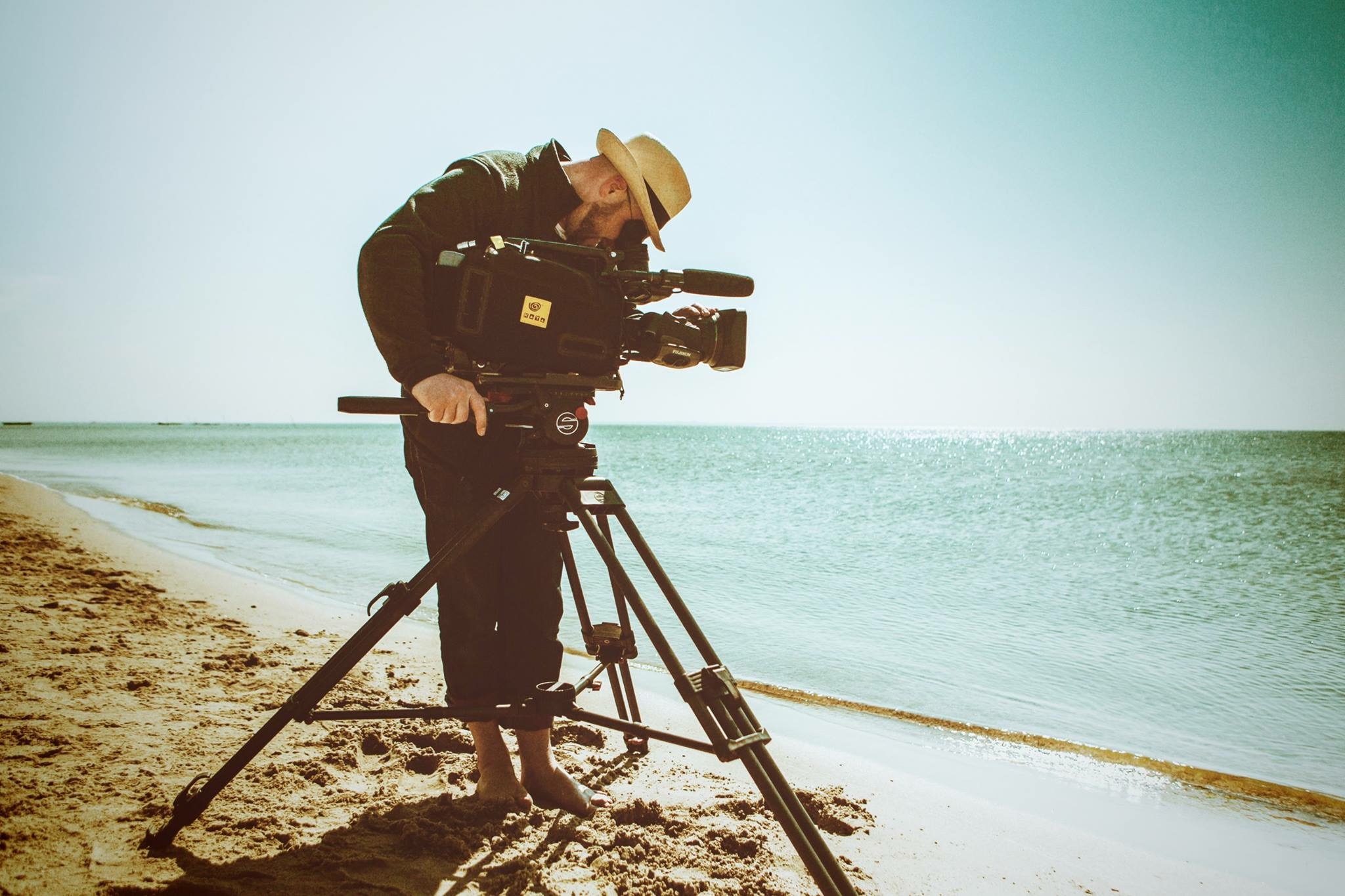 Kameramann mit Strohhut filmt allein am Strand, dahinter das Meer